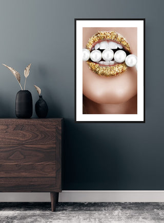 Framed poster of glossy golden lips holding pearls, displayed above a modern wooden dresser in a stylish room.
