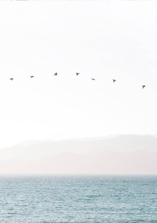 Minimalist artwork of birds flying in a straight line over calm water with soft mountains in the background.