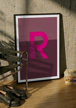 R as in Rebel – Cerise by Quite Nice Prints in a black frame leaning against a concrete wall by a steel chair