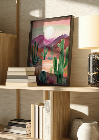 Cactus desert by Ohkimiko in a dark frame on a wooden shelf beside books and a lamp