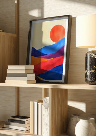 Red sun over colourful hills by Ohkimiko in a dark frame on a wooden shelf beside books and a lamp