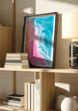 Pink hour on the beach by Ohkimiko in a dark frame on a wooden shelf beside books and a lamp