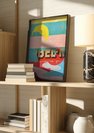 BEL-M by Kareem Rizk in a dark frame on a wooden shelf beside books and a lamp