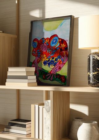 Color frog by Nittaart in a dark frame on a wooden shelf beside books and a lamp