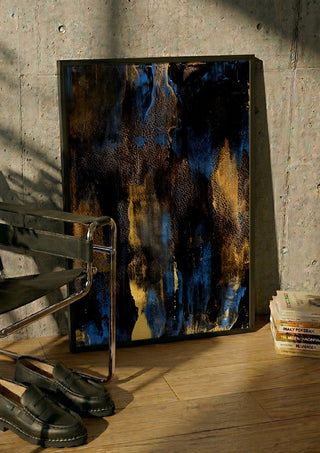 Gold on Blue by Kristof ellertz in a black frame leaning against a concrete wall by a steel chair
