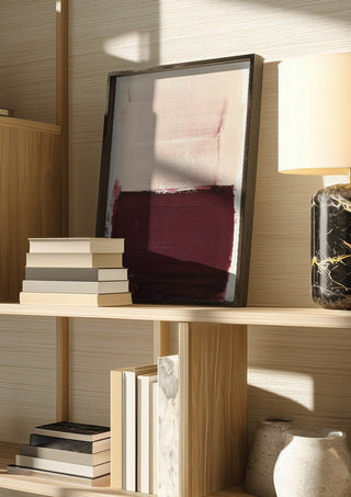 colorfields pale pink warm red by Iris Lehnhardt in a dark frame on a wooden shelf beside books and a lamp