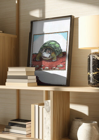 The Reading Tortoise by Waldemar Mandzel in a dark frame on a wooden shelf beside books and a lamp