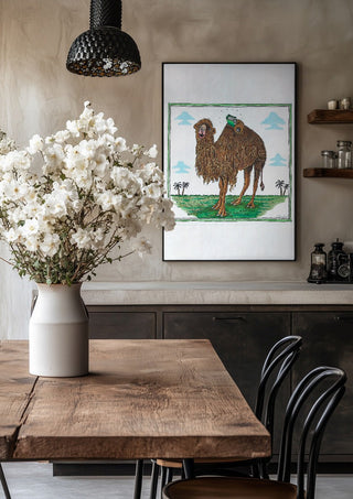 The Camel and the Book by Waldemar Mandzel in a black frame above a rustic table with white flowers