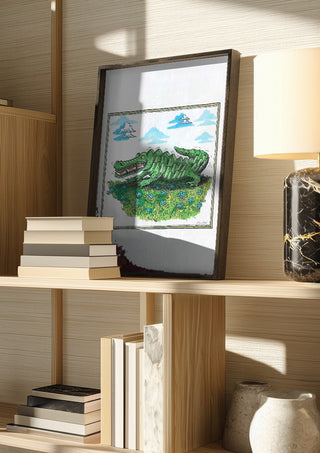 The Crocodile and the Book by Waldemar Mandzel in a dark frame on a wooden shelf beside books and a lamp