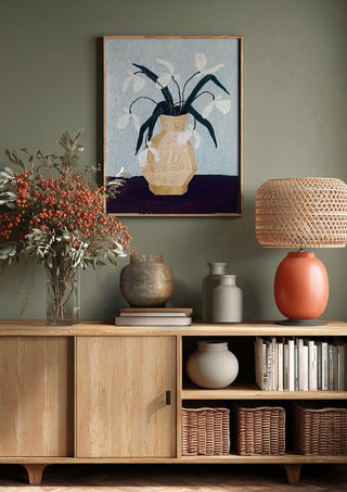 Vita blomster i beige vas by Rebecka Hansson above a wooden sideboard on a sage green wall