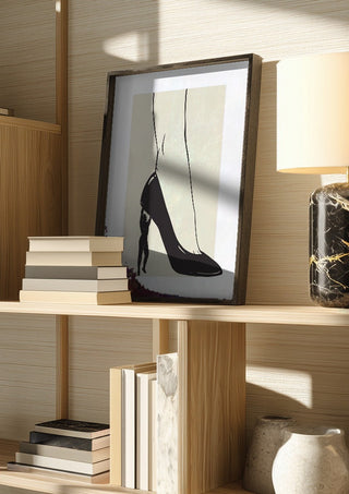 Heavy is the Heel by Nari in a dark frame on a wooden shelf beside books and a lamp