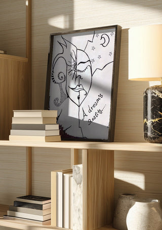 A dreams' reality by A Sleepy Spirit Art in a dark frame on a wooden shelf beside books and a lamp